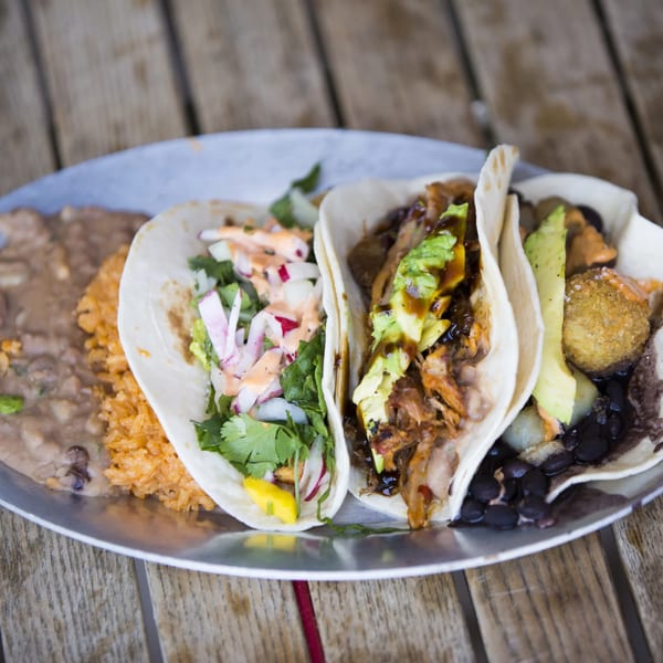 Three tacos with various fillings, rice, and beans on a metal plate on a wooden table.