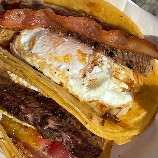 Close-up of a stacked cheeseburger with a fried egg, bacon, beef patties, tomato, and toasted bun.