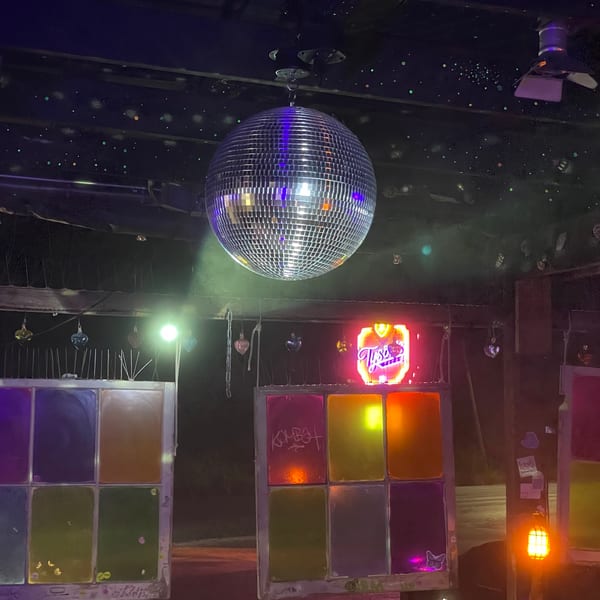 A disco ball hangs from a dark ceiling with neon lights and colorful window panels below.