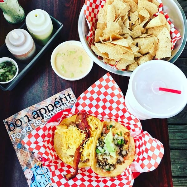Top-down view of tacos, tortilla chips, queso dip, condiments, and a drink served on red-and-white checkered paper.