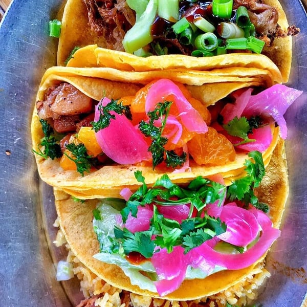 Three colorful tacos with various toppings, including pickled onions, oranges, and green onions, on a metal plate.