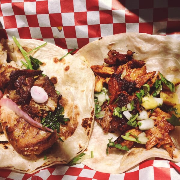 Two tacos, one with chicken and orange, the other with pork and pineapple, on red checkered paper.