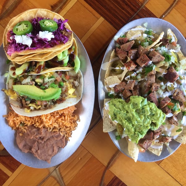 Mexican meal featuring tacos, steak nachos with guacamole, rice, refried beans, avocado, jalapeños, and purple cabbage on plates.