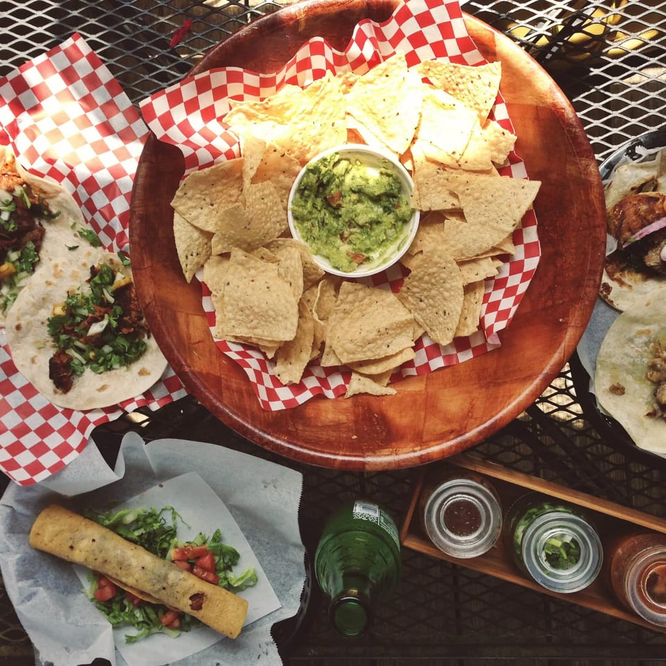Overhead spread of tacos, rolled taquito, tortilla chips with guacamole, sauces, and drinks on a restaurant table.