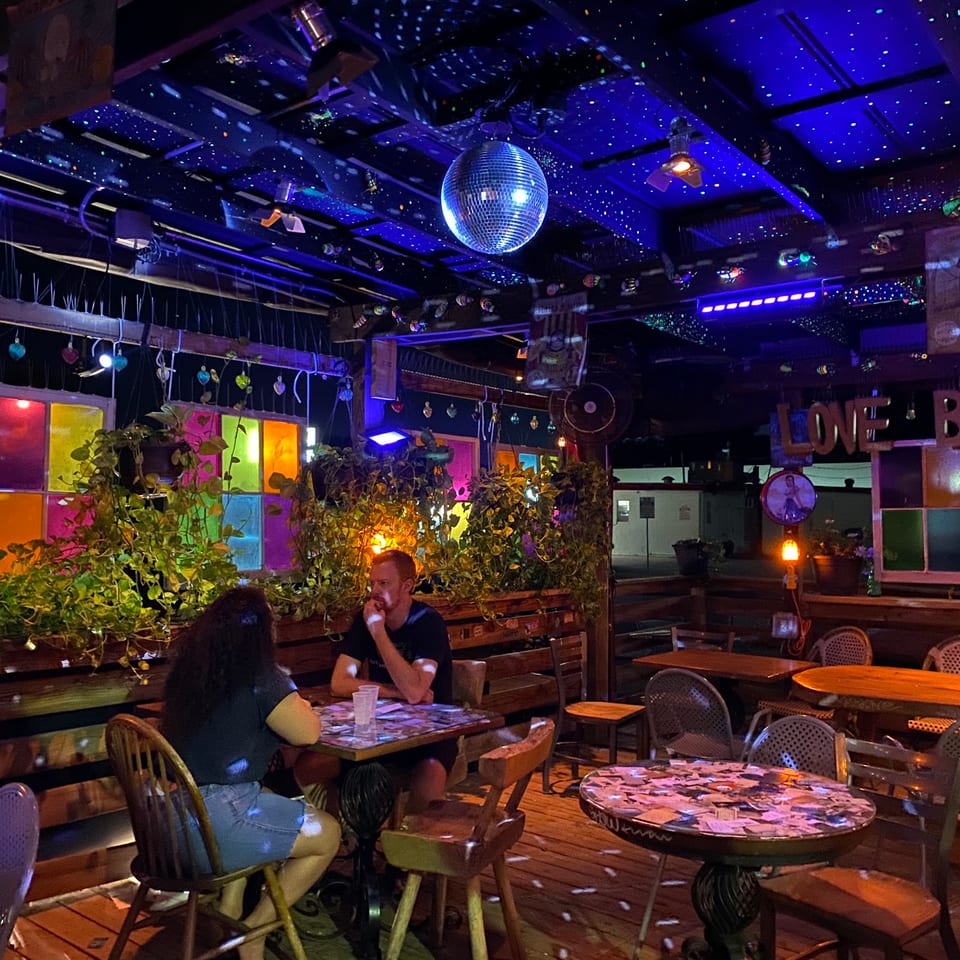A couple sits at a table in a dimly lit bar with a disco ball and colorful windows.