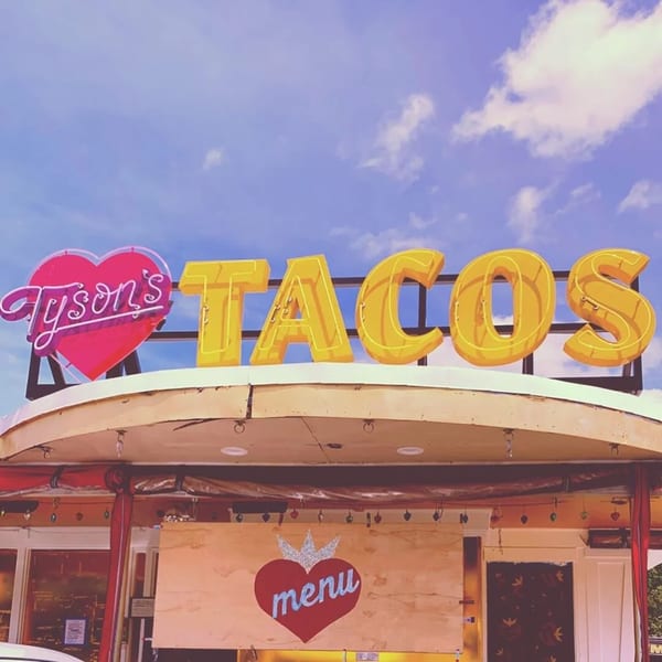 Neon sign for "Tyson's Tacos" with a pink heart and yellow letters against a blue sky.